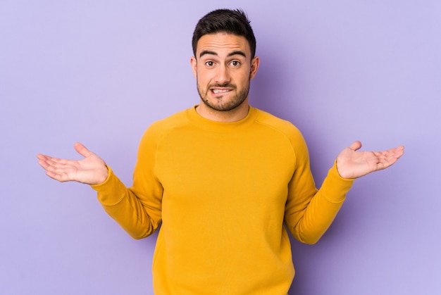 Young caucasian man isolated on purple confused and doubtful shrugging shoulders to hold a copy space.