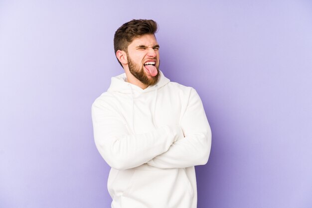 Young caucasian man isolated on purple funny and friendly sticking out tongue.