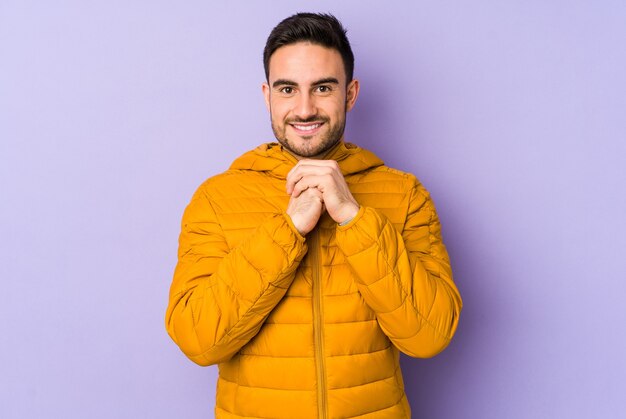 Young caucasian man isolated on purple keeps hands under chin, is looking happily aside.