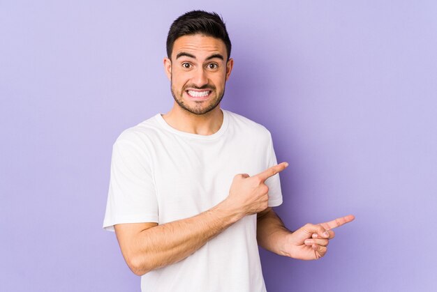 Young caucasian man isolated on purple shocked pointing with index fingers to a copy space.