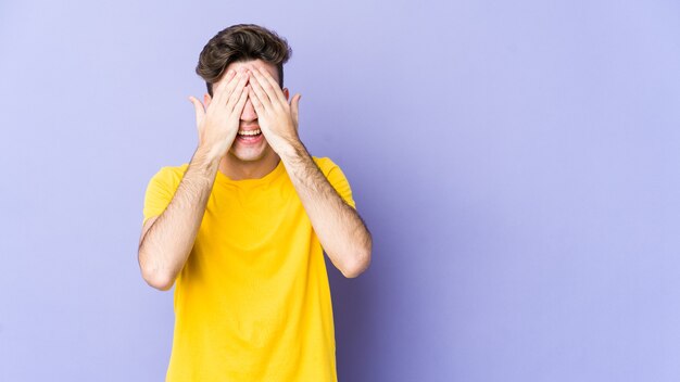 Young caucasian man isolated on purple space covers eyes with hands, smiles broadly waiting for a surprise.