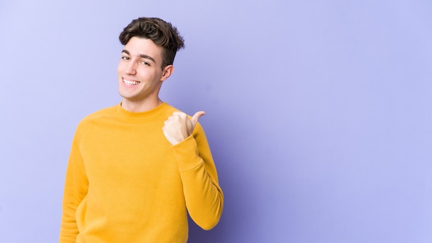 Young caucasian man isolated on purple wall points with thumb finger away, laughing and carefree.