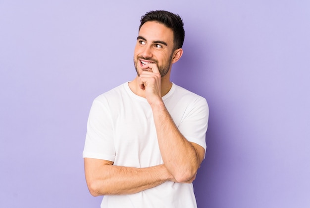 Young caucasian man isolated on purple wall relaxed thinking about something looking at a copy space.