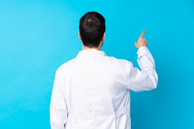 Young caucasian man over isolated wall wearing a doctor gown and pointing back