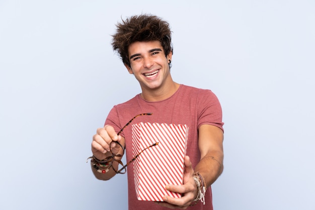 Молодой кавказский человек над изолированной стеной с стеклами и держать большое ведро попкорнов
