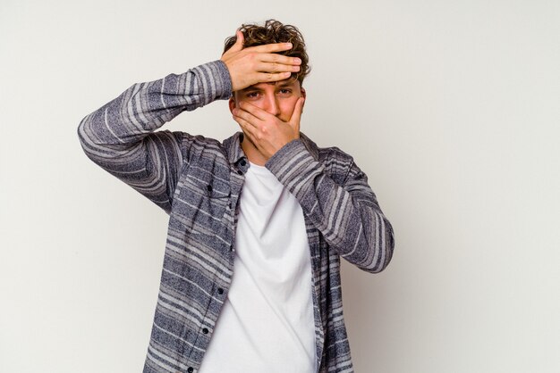 Young caucasian man isolated on white background blink at the camera through fingers, embarrassed covering face.