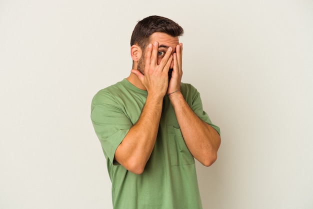 Young caucasian man isolated on white background blink through fingers frightened and nervous.