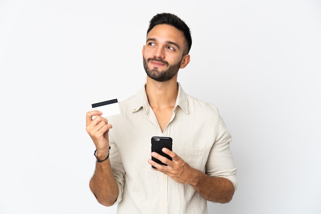 Young caucasian man isolated on white background buying with the mobile with a credit card while thinking