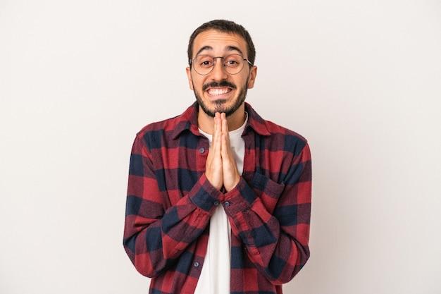 Young caucasian man isolated on white background holding hands in pray near mouth, feels confident.