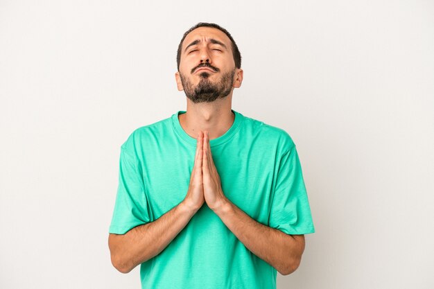 Young caucasian man isolated on white background holding hands in pray near mouth, feels confident.