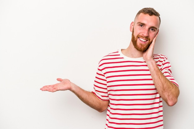 Young caucasian man isolated on white background holds copy space on a palm, keep hand over cheek. Amazed and delighted.