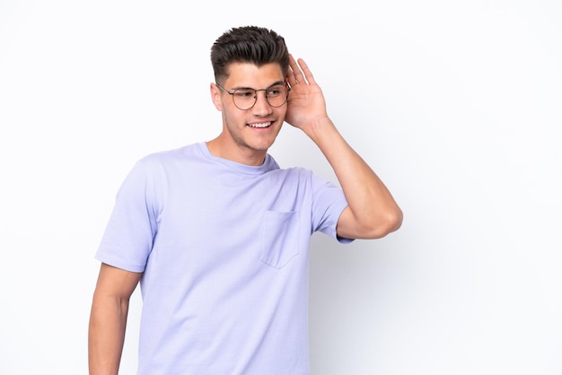 Young caucasian man isolated on white background listening to something by putting hand on the ear