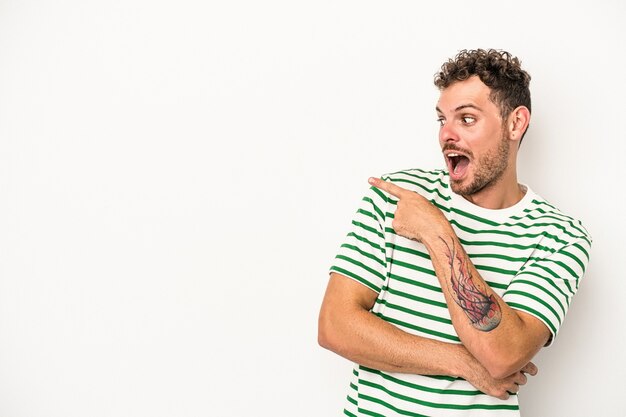 Young caucasian man isolated on white background points with thumb finger away, laughing and carefree.