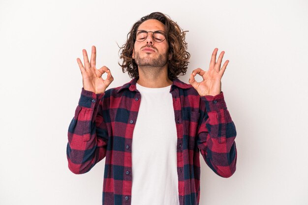 Il giovane uomo caucasico isolato su sfondo bianco si rilassa dopo una dura giornata di lavoro, sta eseguendo yoga.