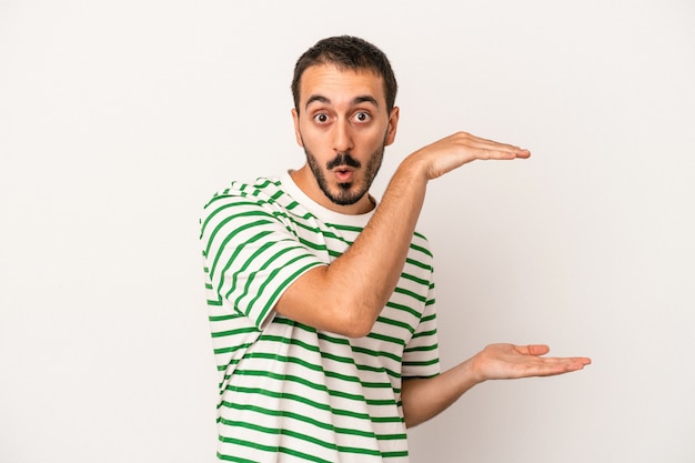 Young caucasian man isolated on white background shocked and amazed holding a copy space between hands.