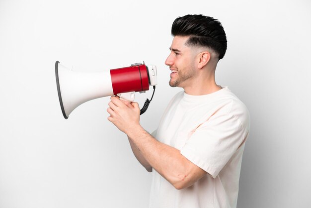 Foto giovane uomo caucasico isolato su sfondo bianco che grida attraverso un megafono per annunciare qualcosa in posizione laterale