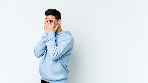 young caucasian man isolated on white space blink through fingers frightened and nervous.