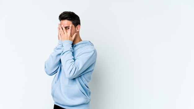 Young caucasian man isolated on white space blink through fingers frightened and nervous.