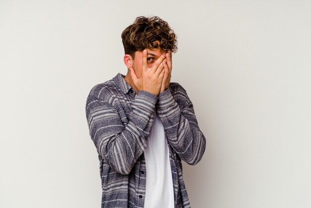 Young caucasian man isolated on white wall blink through fingers frightened and nervous.