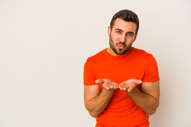 Young caucasian man isolated on white wall folding lips and holding palms to send air kiss.