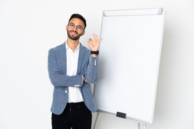 Young caucasian man isolated on white wall giving a presentation on white board and showing ok sign with fingers