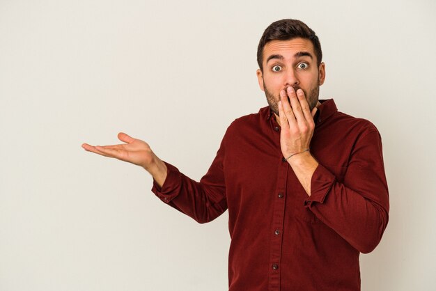 Young caucasian man isolated on white wall impressed holding copy space on palm.