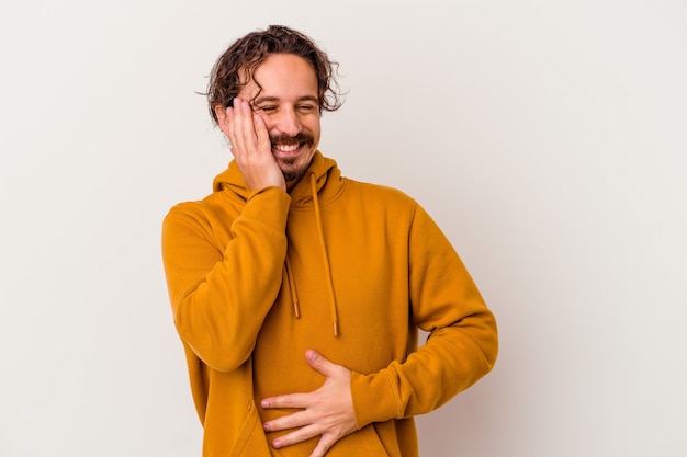 Young caucasian man isolated on white wall laughs happily and has fun keeping hands on stomach.