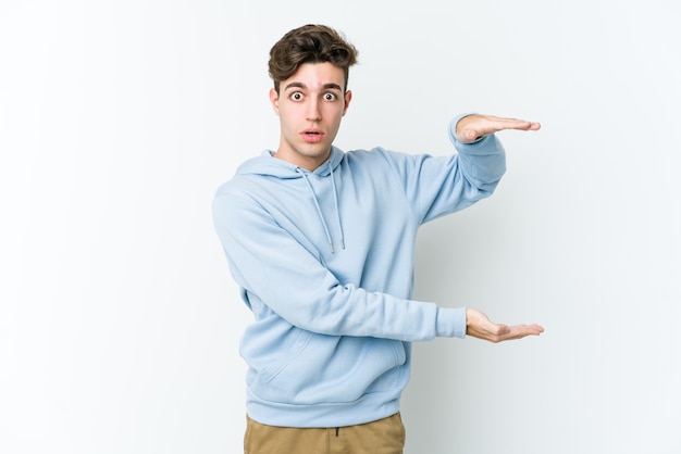 Young caucasian man isolated on white wall shocked and amazed holding a copy space between hands.