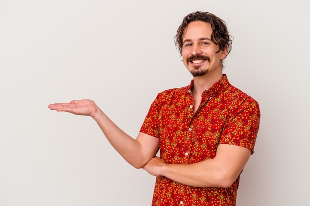 Young caucasian man isolated on white wall showing a copy space on a palm and holding another hand on waist.