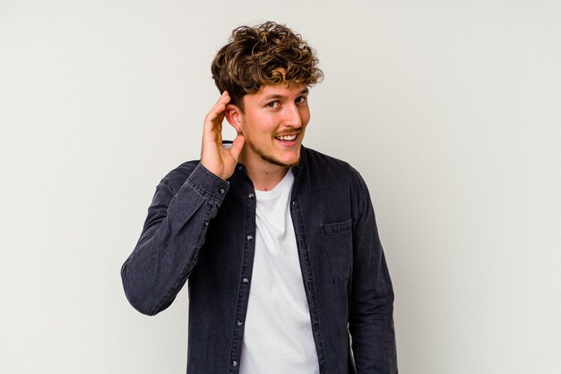 Young caucasian man isolated on white wall trying to listening a gossip.