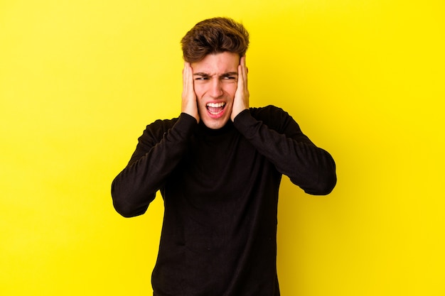 Young caucasian man isolated on yellow background covering ears with hands trying not to hear too loud sound.