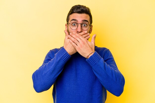 Young caucasian man isolated on yellow background covering mouth with hands looking worried