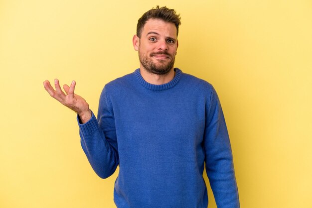 Young caucasian man isolated on yellow background doubting and shrugging shoulders in questioning gesture.