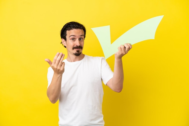 Young caucasian man isolated on yellow background holding a check icon and doing coming gesture