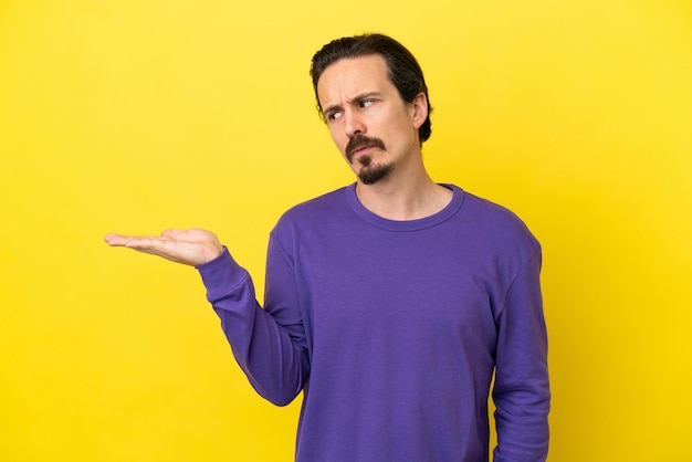 Young caucasian man isolated on yellow background holding copyspace with doubts