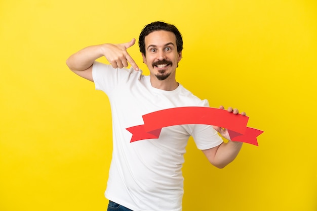 Young caucasian man isolated on yellow background holding an empty placard and pointing it