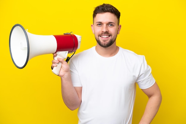 Young caucasian man isolated on yellow background holding a megaphone and smiling