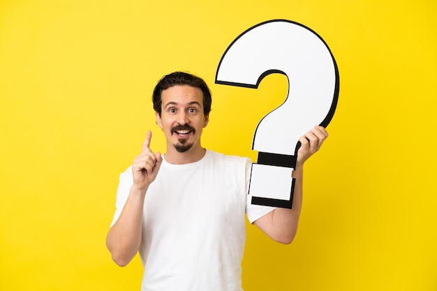 Young caucasian man isolated on yellow background holding a question mark icon