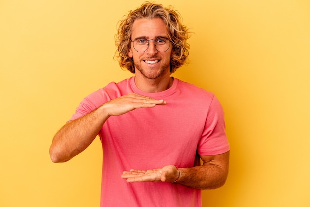 Young caucasian man isolated on yellow background holding something with both hands, product presentation.