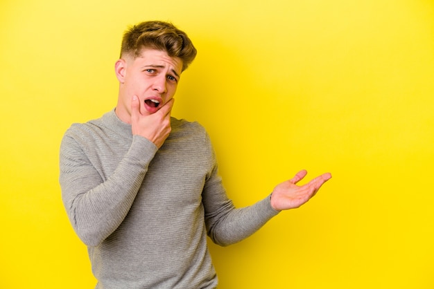 Young caucasian man isolated on yellow background holds copy space on a palm, keep hand over cheek. Amazed and delighted.