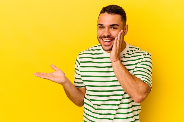 Young caucasian man isolated on yellow background holds copy space on a palm, keep hand over cheek. Amazed and delighted.