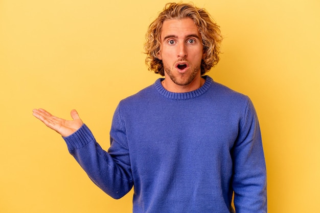 Young caucasian man isolated on yellow background impressed holding copy space on palm.