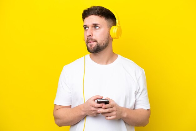 Young caucasian man isolated on yellow background listening music with a mobile and thinking