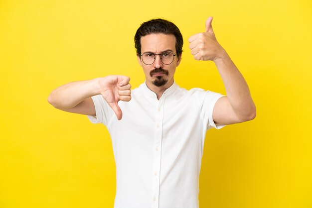 Young caucasian man isolated on yellow background making good-bad sign. Undecided between yes or not