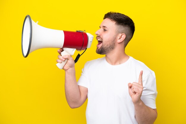 Young caucasian man isolated on yellow background shouting through a megaphone to announce something in lateral position