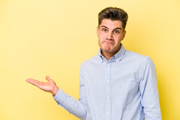 Young caucasian man isolated on yellow background showing a copy space on a palm and holding another hand on waist