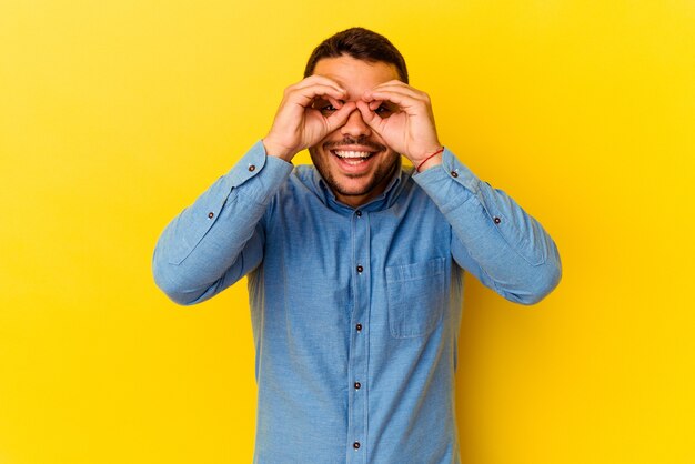 Young caucasian man isolated on yellow background showing okay sign over eyes