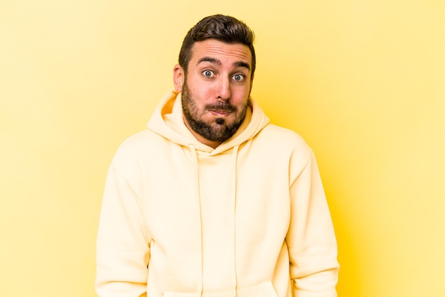 Young caucasian man isolated on yellow background shrugs shoulders and open eyes confused