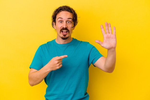 Young caucasian man isolated on yellow background smiling cheerful showing number five with fingers.