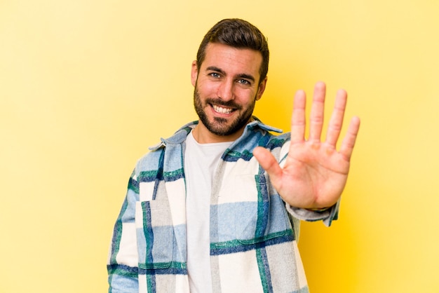 Young caucasian man isolated on yellow background smiling cheerful showing number five with fingers